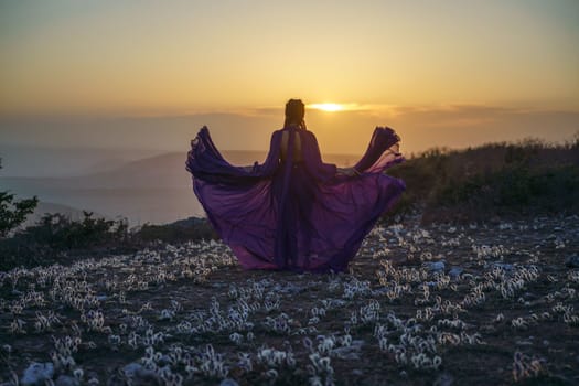 Sunset purple dress woman mountains. Rise of the mystic. sunset over the clouds with a girl in a long purple dress. In the meadow there is a grass dream with purple flowers