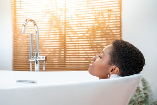 Rest, calm, pleasure, happiness and wellbeing concept. Attractive African woman relaxing in foam bath in beautiful bathroom with plants at home during a sunny morning routine. High quality photo