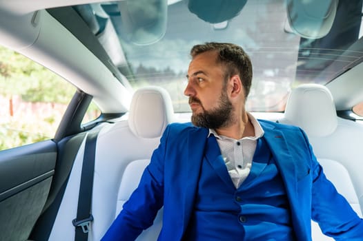 A caucasian man in a blue suit looks out the open window while sitting in the back seat of a car. Business class passenger