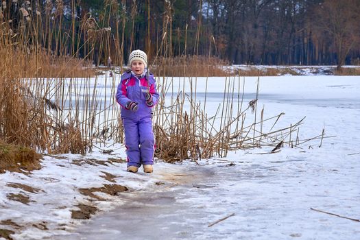 a young human being below the age of puberty or below the legal age of majority. A cute child in a lilac jumpsuit walks on the lake in a winter park.