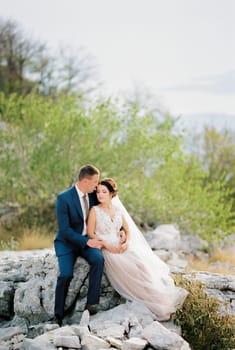 Groom hugs bride in a white dress sitting on the stones. High quality photo