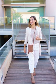 Full body happy adult woman in stylish clothes with smartphone smiling and looking away while walking on path against modern building