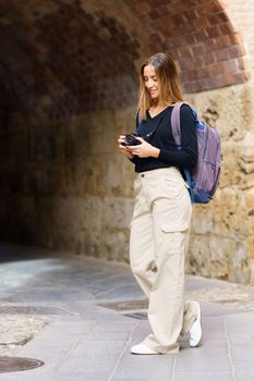 Side view of content young female traveler with long brown hair in casual clothes, and backpack taking photos on camera and smiling while standing near archway in old city during vacation