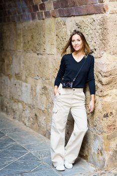 Positive young female tourist with long brown hair and photo camera, in casual clothes smiling and looking at camera while leaning on old stone wall during vacation