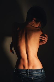 Back view of adult alluring topless woman with short dark hair in jeans standing near wall with crossed arms