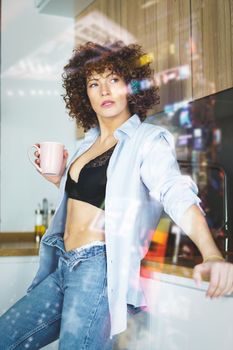 View through glass of adult female in unbuttoned blue shirt and unzipped jeans leaning on counter while having cup of hot beverage looking away