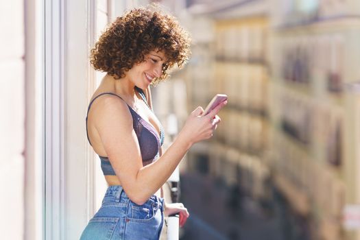 Side view of positive young female in bra and jeans with curly hair smiling while browsing smartphone on sunny balcony