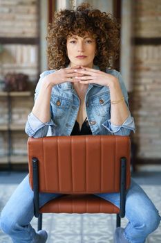 Thoughtful Young female with afro hair in black bra and denim jacket sitting on brown leather chair and touching chin while looking at camera