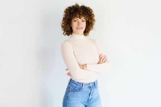 Confident adult curly haired female model wearing beige turtleneck and blue jeans standing with arms crossed against white background smiling at camera