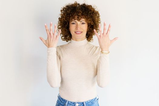 Positive curly haired female showing number ten on fingers while raising arms and looking at camera against white background