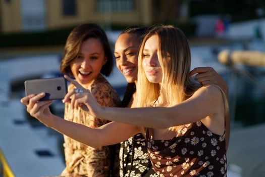 Smiling young multiethnic female friends taking self portrait on cellphone while sitting together on pier against blurred background on summer day