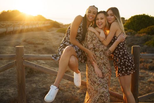 Delighted multiracial girlfriends in summer dresses cuddling near wooden fence on boardwalk on sandy coast at sunset