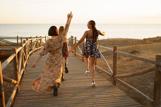 Full body back view of multiracial girlfriends raising arms while running on wooden pier towards endless sea at sunset