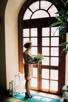 Bride with a bouquet in her hands stands near the arched glass door. High quality photo