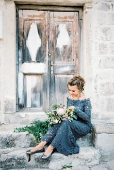 Bride with a bouquet of flowers on her knees sits on the steps of an old building near the door. High quality photo