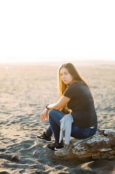 Girl in jeans sits on a snag on a sandy beach. High quality photo