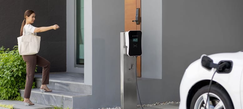 Asian woman lock electric vehicle with remote car key at her garage, EV car recharge battery at home charging station. Alternative clean energy applied in daily life as progressive lifestyle concept.