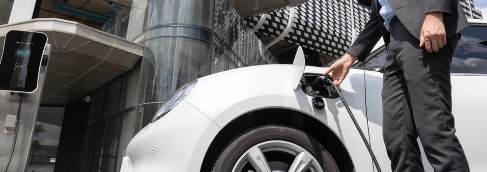 Closeup progressive man holding EV charger plug from public charging station for electric vehicle with background of residential building as concept eco-friendly sustainability energy car concept.