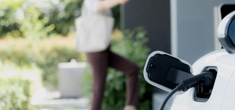 Focus EV charger plugged into EV car at home charging station with blurred background of progressive woman walking in background. Elective vehicle powered by clean energy for eco-friendly concept.