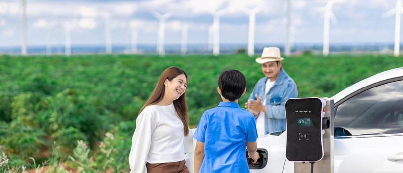 Concept of progressive happy family enjoying their time at wind farm with electric vehicle. Electric vehicle driven by clean renewable energy from wind turbine generator for charging station.