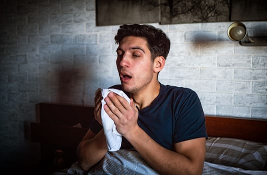 Young man about to sneeze, with handkerchief in his hand, sitting on his bed at home