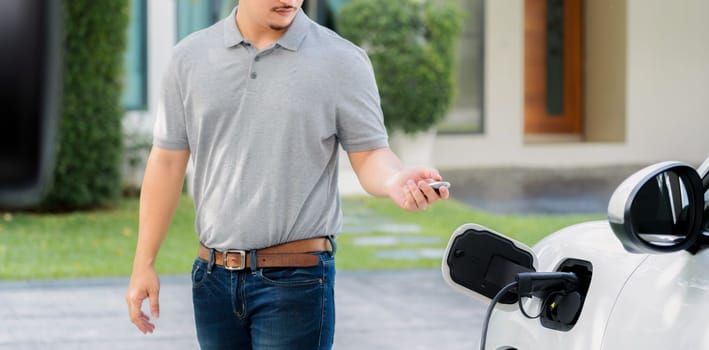 Progressive asian man install cable plug to his electric car with home charging station in the backyard. Concept use of electric vehicles in a progressive lifestyle contributes to clean environment.