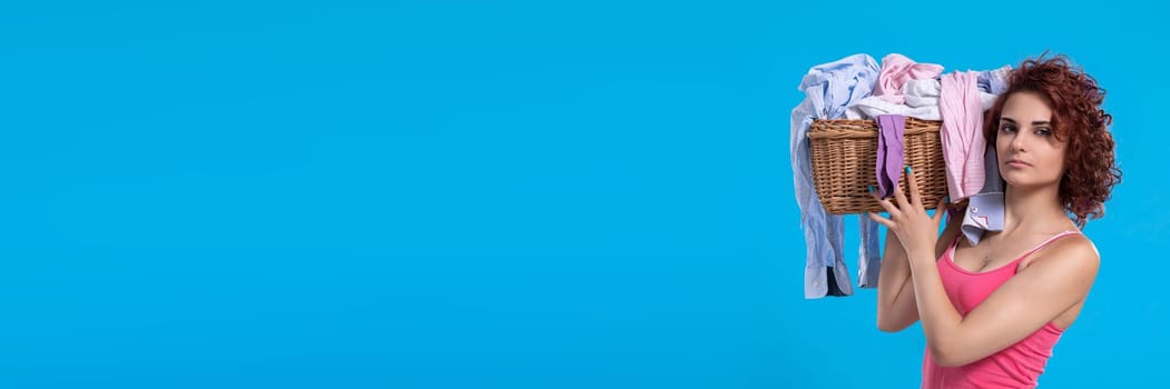 A strong woman holds a heavy wicker basket full of laundry clothes on her shoulder. An attractive girl with dark and curly hair. Panoramic view of girl with empty blue background.