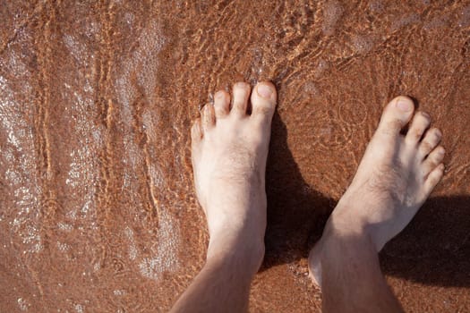 Top view of men's legs in the sea. A man is standing on the sand. An ocean wave washes the shore. Summer holidays.