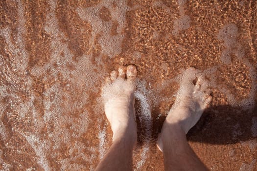 Top view of men's legs in the sea. A man is standing on the sand. An ocean wave washes the shore. Summer holidays.