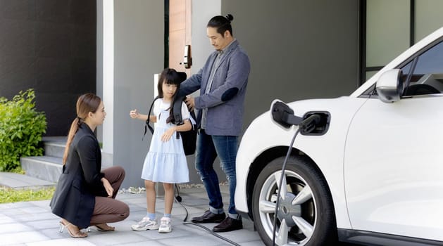 Progressive young parents and daughter with electric vehicle and home charging station. Green and clean energy from electric vehicles for healthy environment. Eco power from renewable source at home.