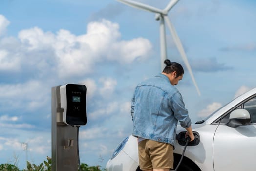Progressive man with his electric car, EV car recharging energy from charging station on green field with wind turbine as concept of future sustainable energy. Electric vehicle with energy generator.