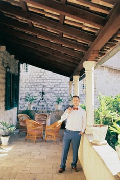 Groom with a jacket on his shoulder stands near the fence of the terrace of an old stone house. High quality photo