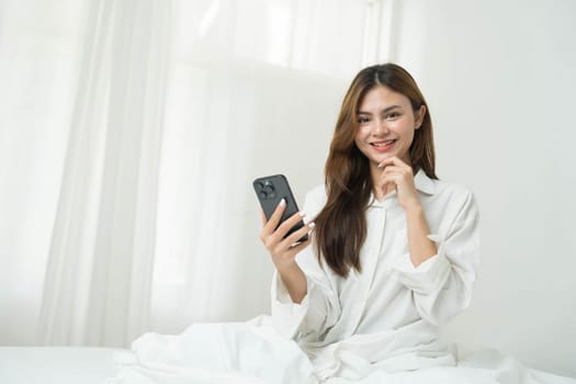 Beautiful young asian woman smiling sitting relax on the bed in the morning, girl using mobile smart phone talking enjoy, communication and social network concept...