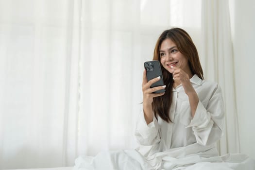 Beautiful young asian woman smiling sitting relax on the bed in the morning, girl using mobile smart phone talking enjoy, communication and social network concept...