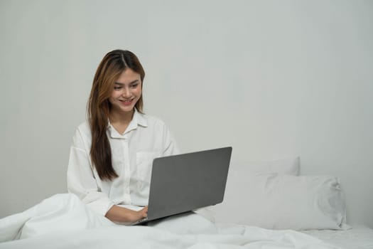 young asian woman using laptop when sitting on a bed. Smart living with communication technology for a better life. girl sitting on at white bedroom space with a clean interior design...