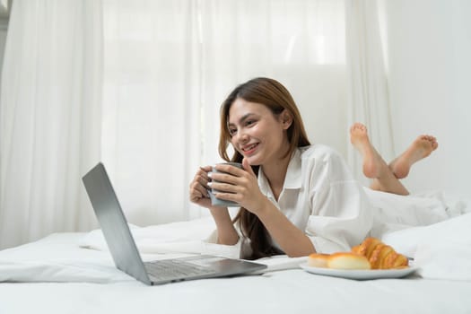 young asian woman using laptop when sitting on a bed. Smart living with communication technology for a better life. girl sitting on at white bedroom space with a clean interior design...