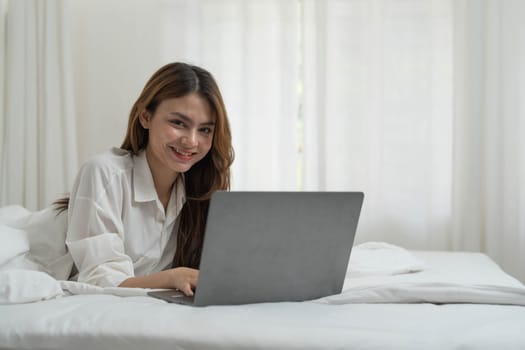 young asian woman using laptop when sitting on a bed. Smart living with communication technology for a better life. girl sitting on at white bedroom space with a clean interior design...
