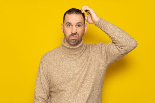 Doubtful Hispanic man thinking, scratching his head trying to find a solution, isolated on a yellow background. He tries to decide where to spend the holidays