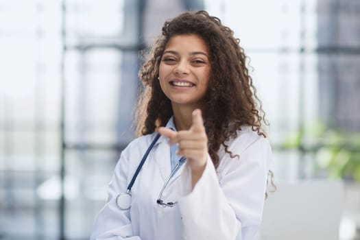 Smiling young female doctor in white coat standing and pointing forward with finger