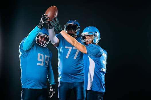 Three American football players raising their hands up holding the ball on a black background