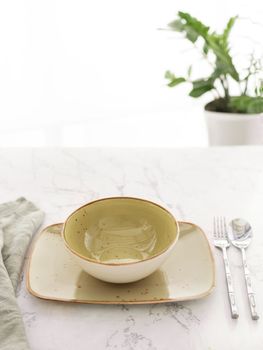 Empty green soup bowl on rectangular plate with tablecloth, spoon and fork on white marble table in front of window