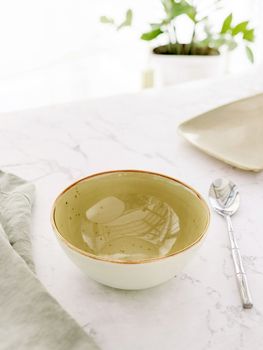 Empty green soup bowl with tablecloth and spoon on white marble table in front of window