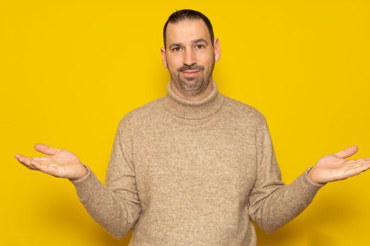 So what, who cares. Unsure and doubtful hispanic man with a beard wearing a beige turtleneck sweater isolated over yellow background. Indecision concept.