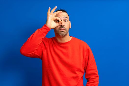 Bearded hispanic man in his 40s wearing a red jumper making the gesture of looking through a telescope by one hand isolated on blue studio background
