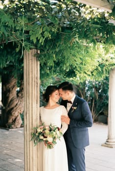 Groom hugs bride from behind by the shoulders near the column in the garden. High quality photo