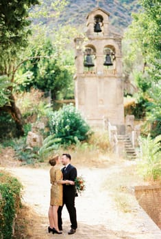 Groom hugs bride near the ancient bell tower in the park. High quality photo