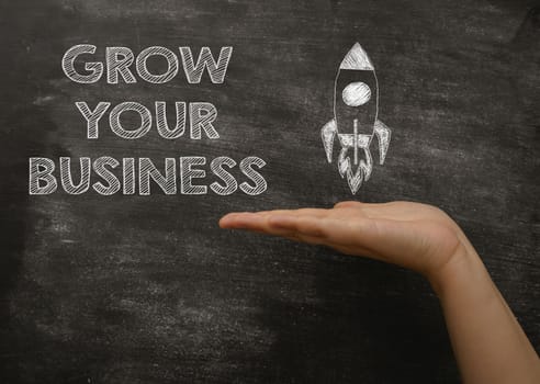 Person palm points to phrase Grow Your Business and rocket picture on blackboard. Step-by-step guide to get more customers to business company
