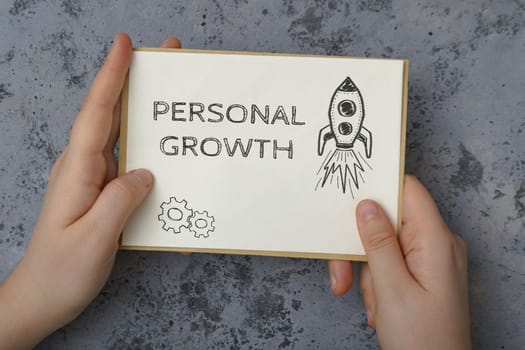 Person hands hold white card with phrase Personal Growth and drawn rocket against concrete background. Human resources and self-motivation in business
