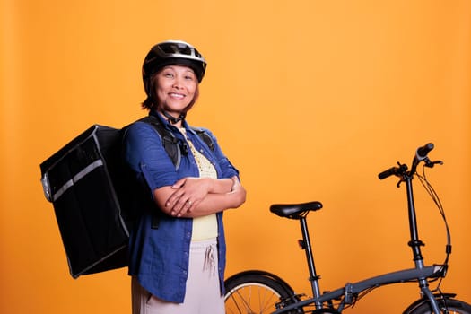 Deliver person carrying food delivery backpack standing beside bike in studio with yellow background. Restaurant worker delivering pizza for lunch. Food transportation service and takeaway concept
