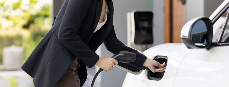 Closeup progressive woman install cable plug to her electric car with home charging station. Concept of the use of electric vehicles in a progressive lifestyle contributes to clean environment.